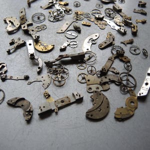 K&ouml;nnte beinhalten: Eine Sammlung antiker Uhrenteile, die auf einer grauen Oberfl&auml;che verstreut sind. Die Ansammlung umfasst kleine Zahnr&auml;der, Hebel und Platten in verschiedenen Formen und Gr&ouml;&szlig;en, mit Silber- und Goldt&ouml;nen. Diese Komponenten deuten auf ein Reparatur- oder Restaurierungsprojekt hin.
