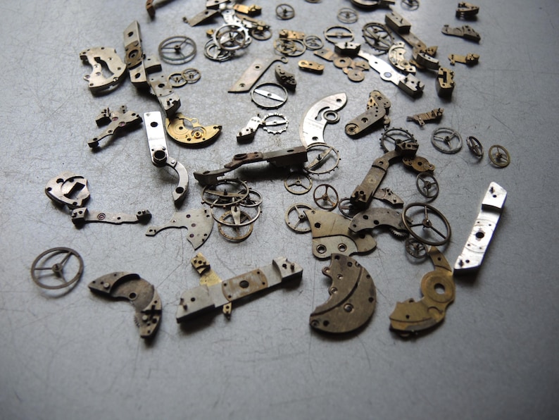 K&ouml;nnte beinhalten: Eine Sammlung antiker Uhrenteile, die auf einer grauen Oberfl&auml;che verstreut sind. Die Ansammlung umfasst kleine Zahnr&auml;der, Hebel und Platten in verschiedenen Formen und Gr&ouml;&szlig;en, mit Silber- und Goldt&ouml;nen. Diese Komponenten deuten auf ein Reparatur- oder Restaurierungsprojekt hin.