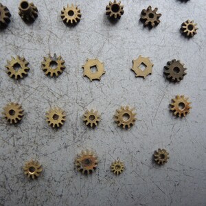 May include: A selection of small brass gears of different sizes, arranged on a grey surface. The gears display various tooth patterns and centre hole designs, highlighting intricate mechanical details. Suitable for watchmaking or steampunk projects.