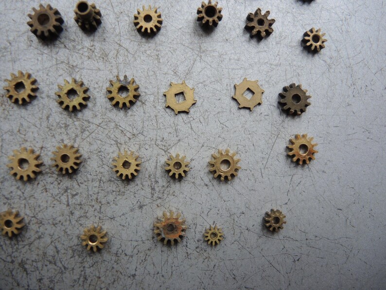May include: A selection of small brass gears of different sizes, arranged on a grey surface. The gears display various tooth patterns and centre hole designs, highlighting intricate mechanical details. Suitable for watchmaking or steampunk projects.