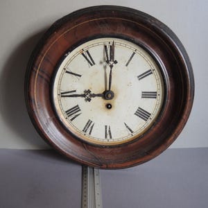 Könnte beinhalten: Runde antike Wanduhr mit einem dunkelbraunen Holzrahmen. Das Zifferblatt ist cremefarben mit schwarzen römischen Ziffern und Zeigern. Die Uhr zeigt ungefähr 9:59 Uhr an.