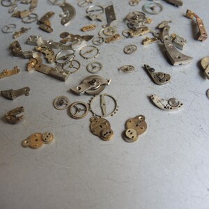 K&ouml;nnte beinhalten: Eine Ansammlung kleiner, zerlegter Uhrwerksteile, die auf einer grauen Oberfl&auml;che verstreut sind. Die Komponenten umfassen verschiedene Zahnr&auml;der, Hebel und Platten in Silber-, Gold- und Bronzet&ouml;nen. Die Teile variieren in Gr&ouml;&szlig;e und Form, was auf eine antike Uhr hindeutet.