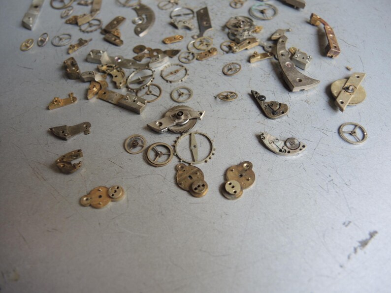 K&ouml;nnte beinhalten: Eine Ansammlung kleiner, zerlegter Uhrwerksteile, die auf einer grauen Oberfl&auml;che verstreut sind. Die Komponenten umfassen verschiedene Zahnr&auml;der, Hebel und Platten in Silber-, Gold- und Bronzet&ouml;nen. Die Teile variieren in Gr&ouml;&szlig;e und Form, was auf eine antike Uhr hindeutet.