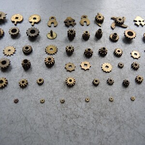 May include: An assortment of small, brass-colored gears of varying sizes and designs. The gears are arranged on a gray surface, showcasing their intricate details and mechanical nature. The collection includes gears with different tooth counts and shapes.