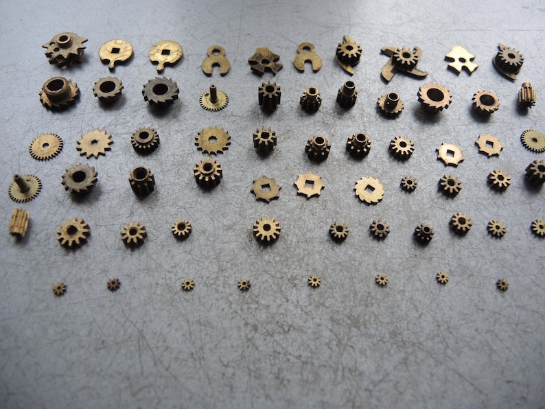 May include: An assortment of small, brass-colored gears of varying sizes and designs. The gears are arranged on a gray surface, showcasing their intricate details and mechanical nature. The collection includes gears with different tooth counts and shapes.