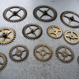 May include: A selection of small, brass and bronze-coloured gears of various sizes. The gears feature a central hole and radiating spokes, with toothed edges. The gears are arranged on a textured, grey surface.