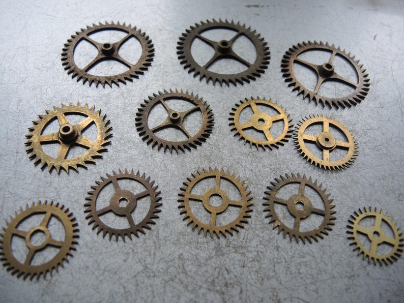 May include: A selection of small, brass and bronze-coloured gears of various sizes. The gears feature a central hole and radiating spokes, with toothed edges. The gears are arranged on a textured, grey surface.