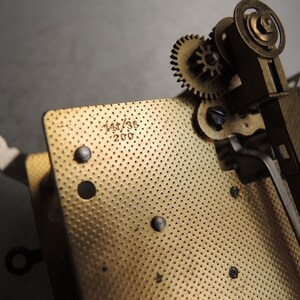 May include: Close-up of a vintage clock mechanism with brass gears and a perforated plate. The plate has the numbers "141/36" and "200" stamped on it. The gears and plate are in a warm, metallic tone.