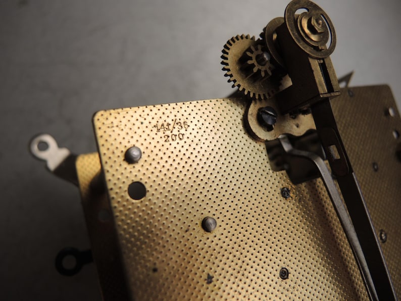 May include: Close-up of a vintage clock mechanism with brass gears and a perforated plate. The plate has the numbers "141/36" and "200" stamped on it. The gears and plate are in a warm, metallic tone.