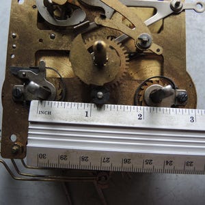 May include: Close-up of a clock mechanism with brass gears and components. A metal ruler is placed across the mechanism, measuring approximately 3 inches. The mechanism has intricate details and various moving parts.