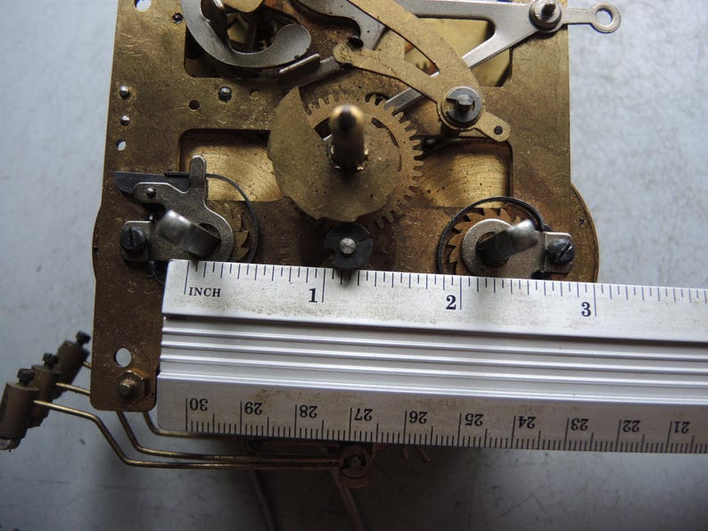 May include: Close-up of a clock mechanism with brass gears and components. A metal ruler is placed across the mechanism, measuring approximately 3 inches. The mechanism has intricate details and various moving parts.