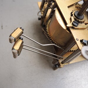 May include: Close-up of a clock mechanism with brass gears and components. Two gold-colored cylindrical weights are attached to thin, silver-colored rods. The intricate design showcases the internal workings of a clock.