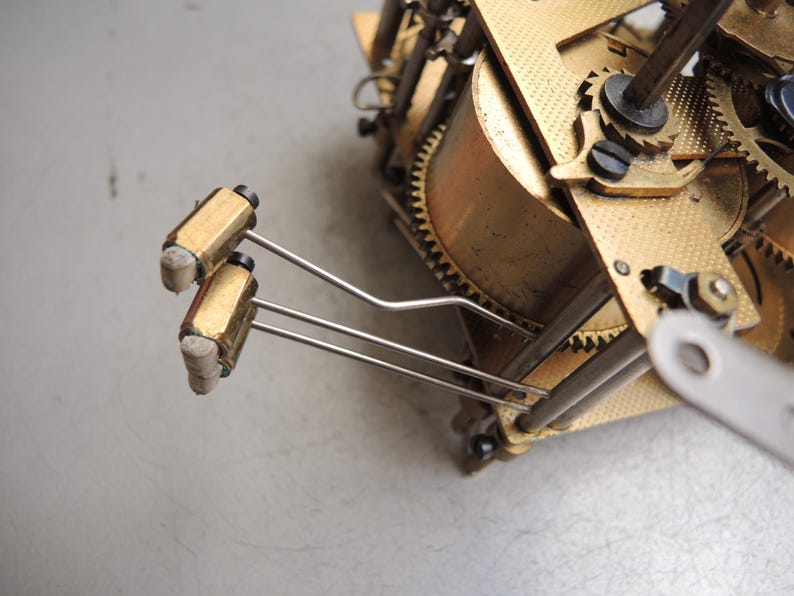 May include: Close-up of a clock mechanism with brass gears and components. Two gold-colored cylindrical weights are attached to thin, silver-colored rods. The intricate design showcases the internal workings of a clock.
