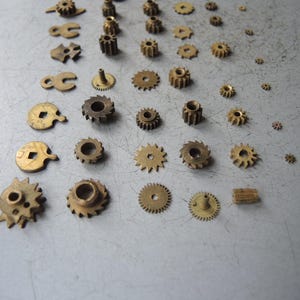 May include: A collection of small, brass-colored gears and watch parts of various sizes and shapes. The components are arranged on a light gray surface, showcasing the intricate details of each piece. Some gears have teeth, while others have holes or unique designs.