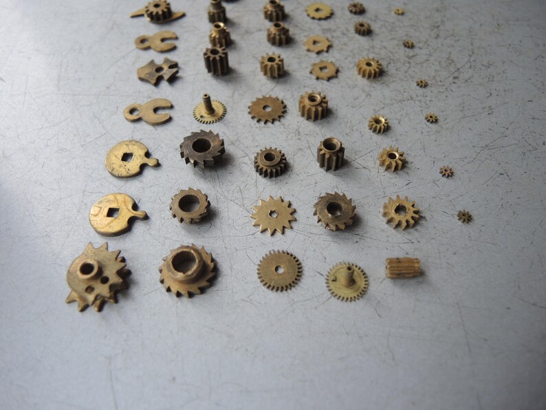 May include: A collection of small, brass-colored gears and watch parts of various sizes and shapes. The components are arranged on a light gray surface, showcasing the intricate details of each piece. Some gears have teeth, while others have holes or unique designs.