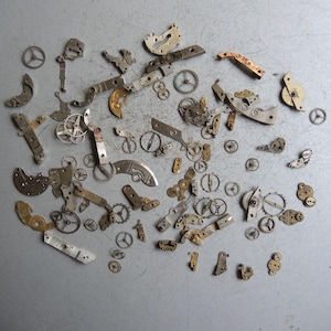 K&ouml;nnte beinhalten: Eine Sammlung antiker Uhrwerksteile, darunter kleine Zahnr&auml;der, Hebel und Platten, auf einer grauen Oberfl&auml;che angeordnet. Die Komponenten sind haupts&auml;chlich metallisch, mit Silber-, Gold- und Bronzet&ouml;nen, was auf Alter und Verwendung in Zeitmessmechanismen hindeutet.