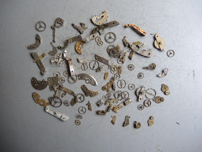 K&ouml;nnte beinhalten: Eine Sammlung antiker Uhrwerksteile, darunter kleine Zahnr&auml;der, Hebel und Platten, auf einer grauen Oberfl&auml;che angeordnet. Die Komponenten sind haupts&auml;chlich metallisch, mit Silber-, Gold- und Bronzet&ouml;nen, was auf Alter und Verwendung in Zeitmessmechanismen hindeutet.