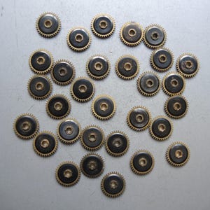 May include: A collection of small, circular watch gears. Each gear has a dark center, a gold-colored toothed outer ring, and a central hole. The gears are scattered across a light gray surface, suggesting a repair or parts supply.