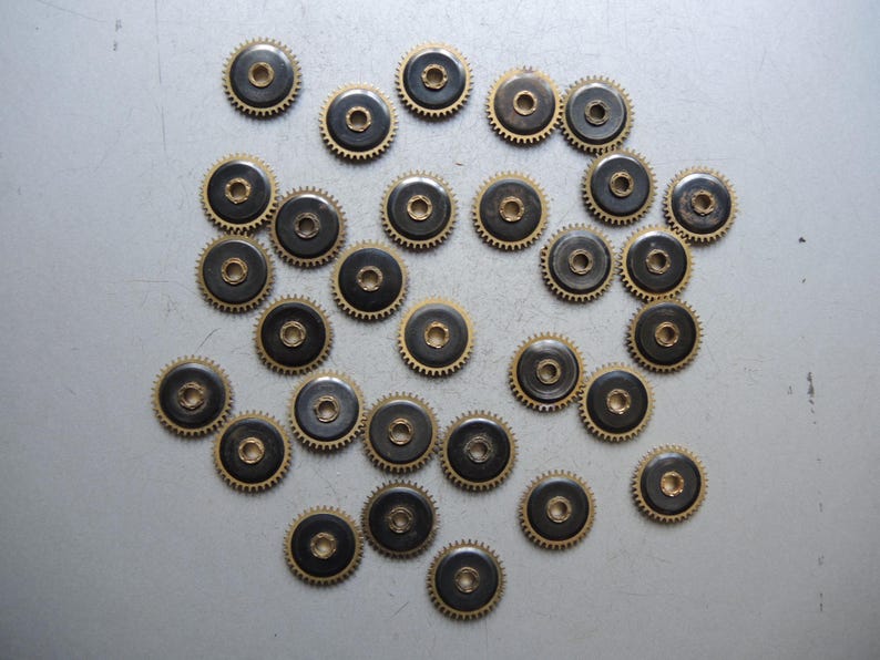 May include: A collection of small, circular watch gears. Each gear has a dark center, a gold-colored toothed outer ring, and a central hole. The gears are scattered across a light gray surface, suggesting a repair or parts supply.