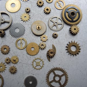 May include: A collection of various sized brass and silver-colored gears scattered on a gray surface. The gears have different designs, including circular, spoked, and toothed variations, suggesting a mechanical or steampunk theme.