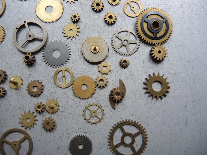 May include: A collection of various sized brass and silver-colored gears scattered on a gray surface. The gears have different designs, including circular, spoked, and toothed variations, suggesting a mechanical or steampunk theme.