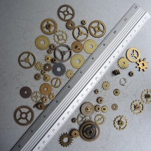 May include: A collection of various sized brass and silver gears, along with a silver ruler, are scattered on a gray surface. The gears range in size and design, showcasing intricate details. The ruler measures up to 12 inches.