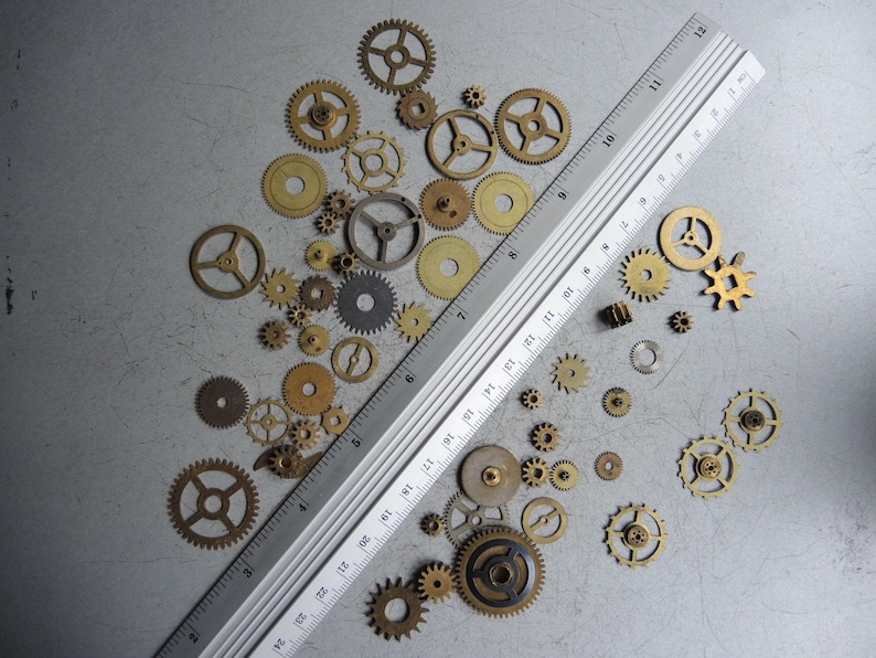 May include: A collection of various sized brass and silver gears, along with a silver ruler, are scattered on a gray surface. The gears range in size and design, showcasing intricate details. The ruler measures up to 12 inches.