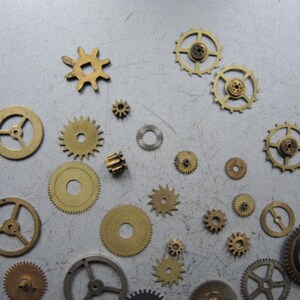 May include: A collection of various sized brass gears scattered on a gray surface. The gears have different tooth counts and designs, with some having spokes and others solid centers. The gears are of different sizes, from small to large.