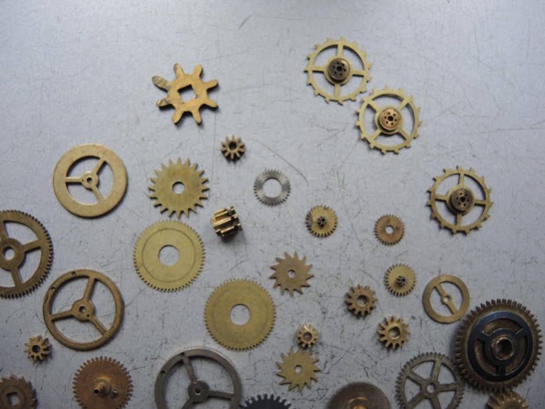 May include: A collection of various sized brass gears scattered on a gray surface. The gears have different tooth counts and designs, with some having spokes and others solid centers. The gears are of different sizes, from small to large.