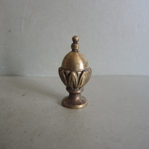 May include: A small, antique brass finial with a decorative, urn-like design. The finial features a rounded top with a small knob and a base with a floral pattern. The piece is likely used for furniture or decorative purposes.