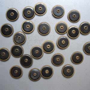 May include: A collection of small, circular clock gears. Each gear has a dark center, a brass-colored toothed outer ring, and a central hole. The gears are scattered across a light gray surface.