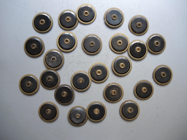 May include: A collection of small, circular clock gears. Each gear has a dark center, a brass-colored toothed outer ring, and a central hole. The gears are scattered across a light gray surface.