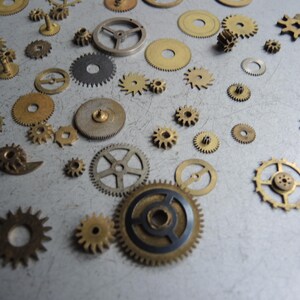 May include: A collection of various sized watch gears in brass, silver, and black. The gears are scattered across a light gray surface, showcasing different shapes and tooth designs. Some gears have a circular design, while others have a star-like appearance.