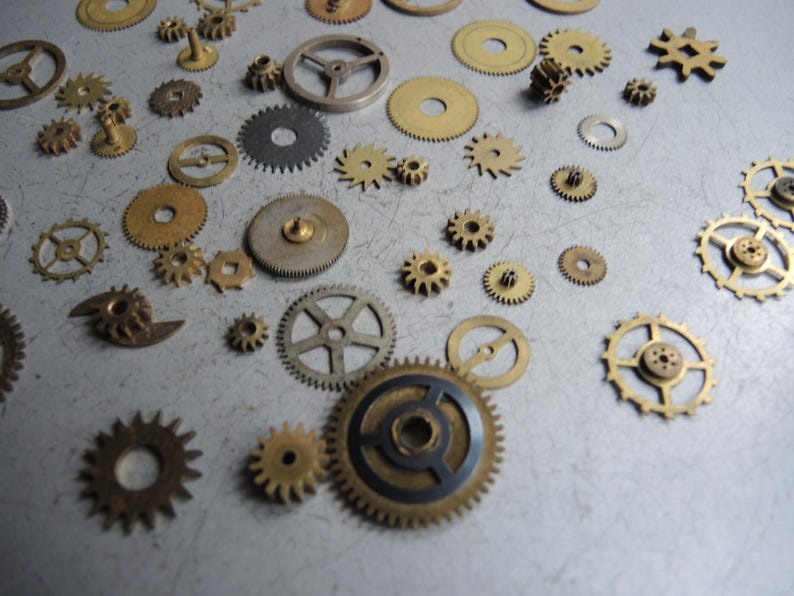May include: A collection of various sized watch gears in brass, silver, and black. The gears are scattered across a light gray surface, showcasing different shapes and tooth designs. Some gears have a circular design, while others have a star-like appearance.