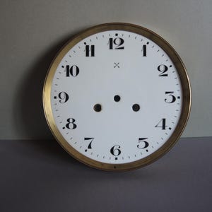 May include: A circular clock face with a white dial and black numerals. The clock face has a gold-coloured rim and two small holes near the centre. The numbers are arranged in a circular pattern, with the number 12 at the top.