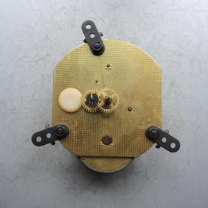 Op de afbeelding: Een vintage uurwerk met een messingkleurige wijzerplaat en zichtbare tandwielen. Het uurwerk heeft drie zwarte montagebeugels en een wit rond onderdeel. De wijzerplaat heeft een gestructureerd, gestippeld patroon.