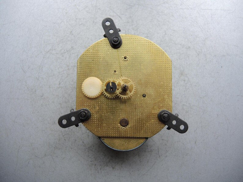 Op de afbeelding: Een vintage uurwerk met een messingkleurige wijzerplaat en zichtbare tandwielen. Het uurwerk heeft drie zwarte montagebeugels en een wit rond onderdeel. De wijzerplaat heeft een gestructureerd, gestippeld patroon.