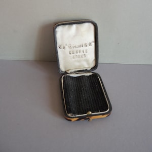 May include: Open antique watch case with a black interior and a white satin lining. The case is rectangular with a hinged lid and a decorative tortoiseshell pattern on the edges. The text "Ch. Tissot & Cie GENEVE 43043" is visible inside the lid.