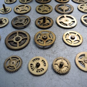 May include: A collection of small, brass-colored gears of varying sizes. The gears have intricate designs with teeth around the edges and central hubs. Some gears have small posts extending from their centers. The gears are arranged on a light gray surface.