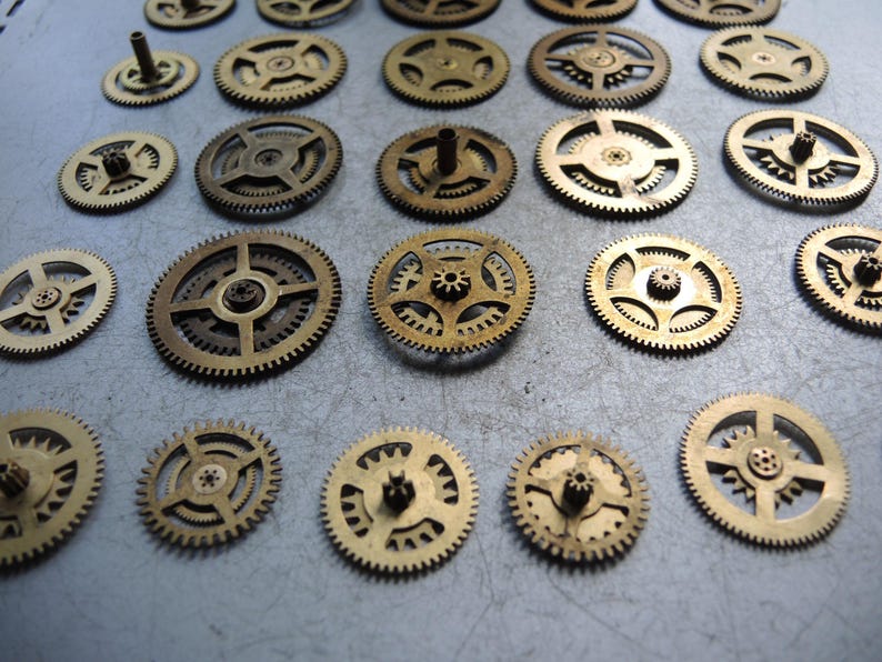 May include: A collection of small, brass-colored gears of varying sizes. The gears have intricate designs with teeth around the edges and central hubs. Some gears have small posts extending from their centers. The gears are arranged on a light gray surface.
