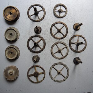 May include: A collection of vintage watch gears and components in various sizes and shapes. The gears are made of brass and have a worn, antique appearance. Some gears have spokes, while others are solid.