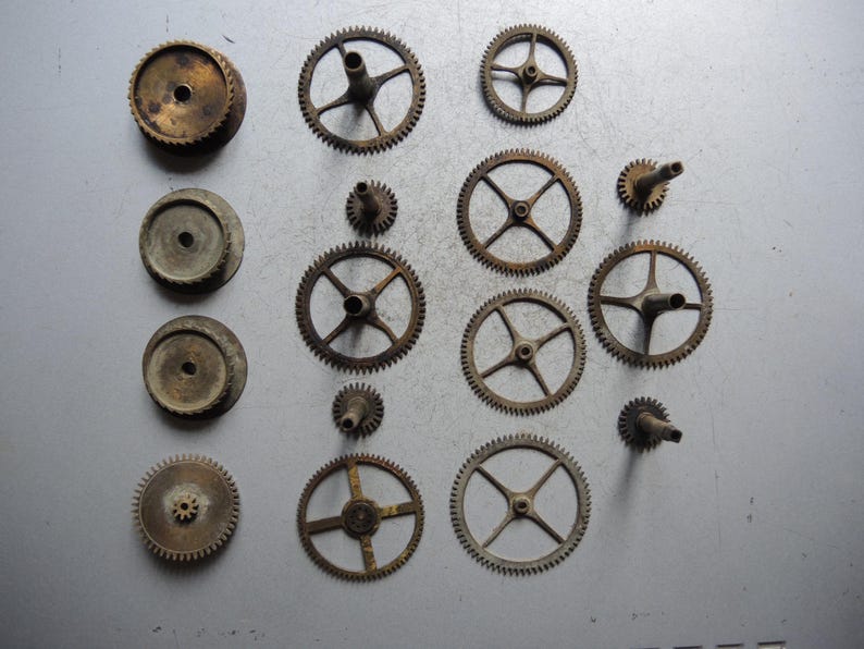 May include: A collection of vintage watch gears and components in various sizes and shapes. The gears are made of brass and have a worn, antique appearance. Some gears have spokes, while others are solid.