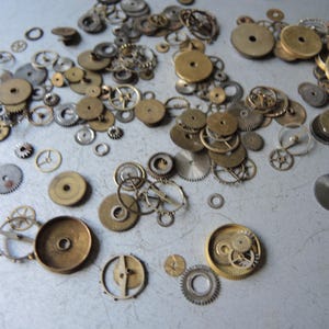 May include: A collection of vintage watch parts, including various sizes of brass and silver-toned gears, wheels, and cogs. The components are scattered across a light gray surface, showcasing their intricate designs and textures.