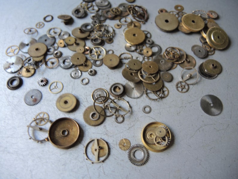 May include: A collection of vintage watch parts, including various sizes of brass and silver-toned gears, wheels, and cogs. The components are scattered across a light gray surface, showcasing their intricate designs and textures.