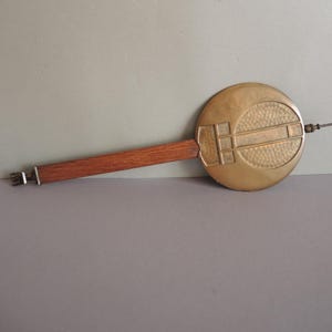 May include: A vintage clock pendulum with a round, brass-colored face and a textured surface. The pendulum has a wooden handle and a metal rod extending from the face. The design includes geometric shapes and lines.