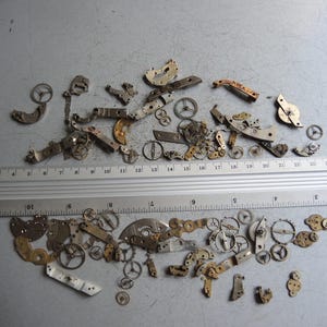 K&ouml;nnte beinhalten: Eine Sammlung kleiner Uhrenteile, darunter Zahnr&auml;der, Hebel und Platten, die auf einer grauen Oberfl&auml;che verstreut sind. Ein Lineal, das in Zentimetern markiert ist, liegt neben den Teilen.