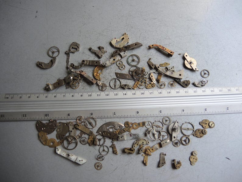 K&ouml;nnte beinhalten: Eine Sammlung kleiner Uhrenteile, darunter Zahnr&auml;der, Hebel und Platten, die auf einer grauen Oberfl&auml;che verstreut sind. Ein Lineal, das in Zentimetern markiert ist, liegt neben den Teilen.