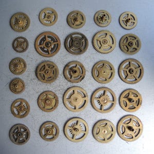 May include: A collection of small, antique brass gears of various sizes. The gears have intricate teeth and central holes, arranged on a gray surface. The gears are likely from clocks or other mechanical devices.