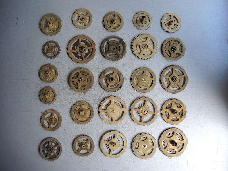 May include: A collection of small, antique brass gears of various sizes. The gears have intricate teeth and central holes, arranged on a gray surface. The gears are likely from clocks or other mechanical devices.