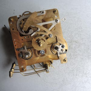 May include: Close-up of a vintage clock movement. The brass-colored mechanism features numerous gears, levers, and springs, mounted on a rectangular plate. The intricate design showcases the internal workings of a clock.